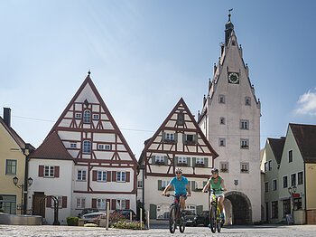 Stadtradeln Monheim 2021 Zwei Personen fahren mit Fahrrädern auf einem gepflasterten Platz, der Monheimer Altstadt. Im Hintergrund Fachwerkhäuser und ein Turm mit einem Torbogen und einer Uhr.