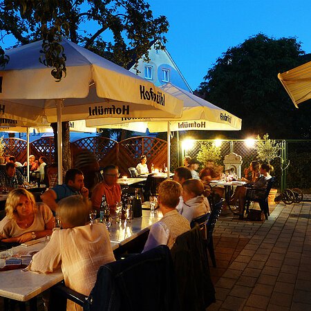 Pizzeria Ristorante Romana - große Terrasse für schöne Stunden im Freien Menschen sitzen abends in einem Biergarten unter großen Sonnenschirmen, umgeben von Holzzäunen und Bäumen.