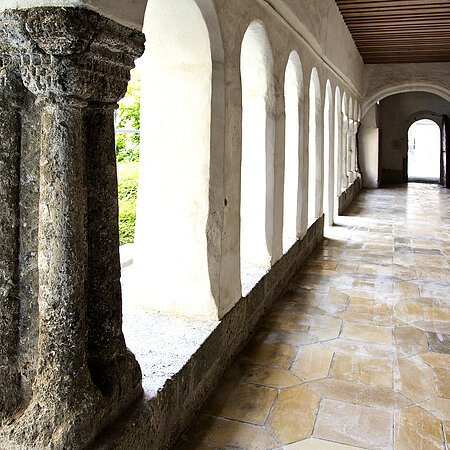 Kreuzgang Monheim Steinbogen-Gang mit Säulen, der Kreuzgang des ehemaligen Klosters in Monheim, führt zu einer Tür, links grüne Büsche sichtbar. Entlang des Gangs alter Bodenbelag.