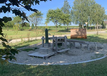 Dorferneuerung Flotzheim Wasserspielplatz Rund gestalteter Wasserspielplatz mit Holz-Wasserpumpe und Sandbereich, umgeben von einer Rasenfläche. Im Hintergrund Bäume und blauer Himmel.