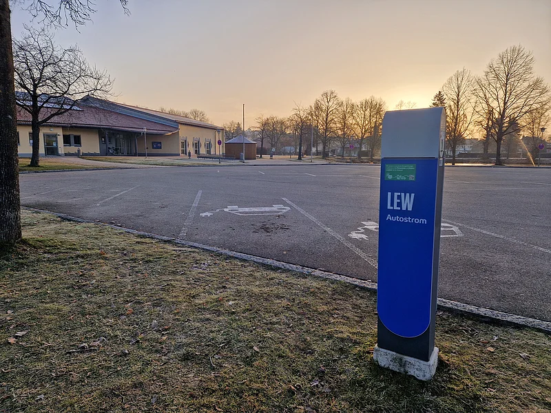 Ladesäule für E-Autos an der Stadthalle Monheim Blaue Ladestation für Elektroautos mit der Aufschrift LEW, auf einem leeren gepflastertem Parkplatz vor einem orangem Gebäude im Hintergrund bei Sonnenaufgang.