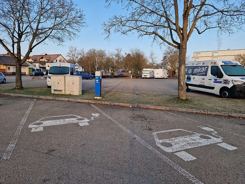 Ladesäule für E-Autos an der Stadthalle Monheim Parkplätze mit Symbolen für Elektroautos auf dem Asphalt und einer dahinterstehender Ladestation in blau, im Freien bei Tageslicht. Im Hintergrund ein Wohnmobilstellplatz mit Parkflächen und mit verschiedenen Fahrzeugen drauf.