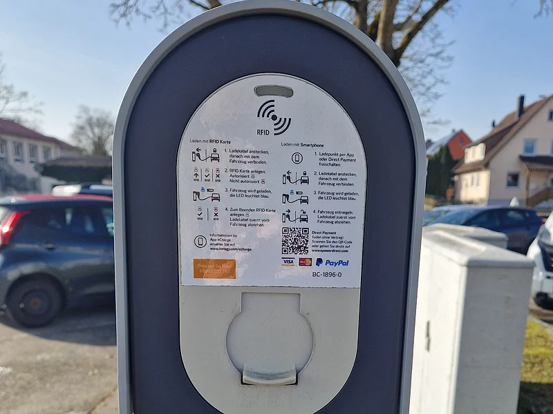 Anschluss der Ladesäule an der Stadthalle Monheim Ladesäule für Elektrofahrzeuge stehend auf einer Grünfläche bei Tageslicht, mit Bedienungsanleitung und Förderhinweis des Bundesministeriums für Verkehr in Wohngebiet.
