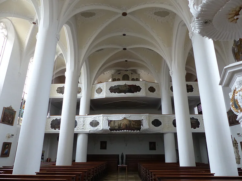 Stadtpfarrkirche St. Walburga Monheim - Blick auf die Empore Innenansicht einer Kirche, der Stadtpfarrkirche St. Walburga in Monheim, mit hohen weißen Säulen, Gewölbedecke, Emporen mit Verzierungen und Holzbänken.