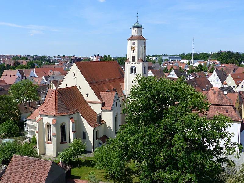 Stadtpfarrkirche St. Walburga Monheim Luftbild der Stadtpfarrkirche mit Turm in einer Stadtlandschaft, umgeben von Häusern mit roten Dächern und Bäumen. Im Hintergrund blauer Himmel.