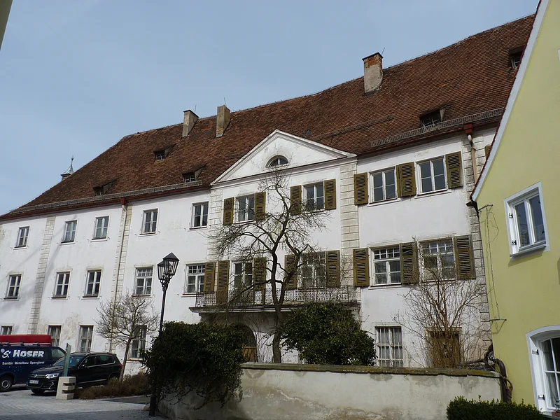 Ehemaliges Schloss / Amtsgericht Monheim Altes, historisches, mehrstöckiges Gebäude mit braunem Ziegeldach, weißen Wänden und grünen Fensterläden bei Tageslicht. Vor dem Gebäude sind Bäume, parkende Autos und eine Straßenlaterne zu sehen. Rechts im Bild Stück einer weißer Mauer und ein grünes Gebäude zu sehen.