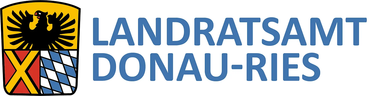 Wappen des Landratsamts DONAU-RIES: auf dem Wappen oben ein schwarzer Adler auf gelbem Hintergrund. darunter links ein gelbes Kreuz auf rotem Hintergrund und rechts davon bayerisches Rautenmuster in weiß-blau. Rechts neben dem Wappen der Text: Landratsamt Donau-Ries.