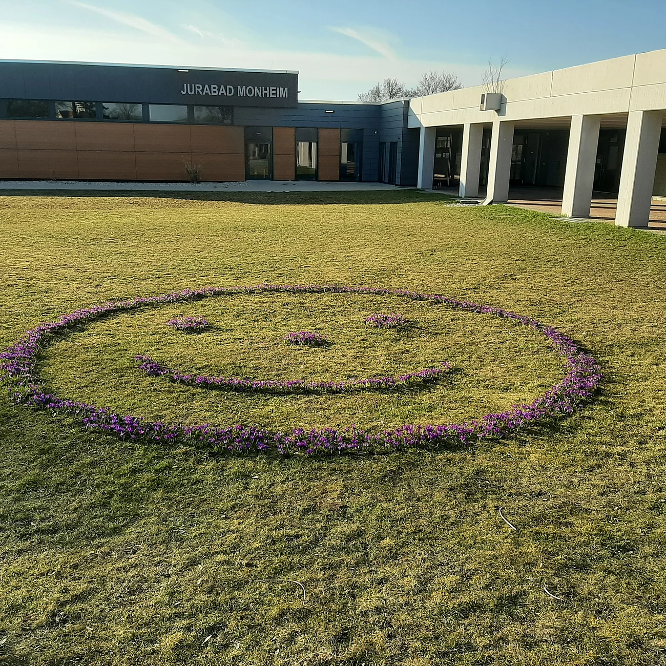 Blumenbeet in Form eines Smileys auf einer Rasenfläche vor einem modernen Gebäude, in grau mit einer Holzfassade. Darauf die Aufschrift "Jurabad Monheim".