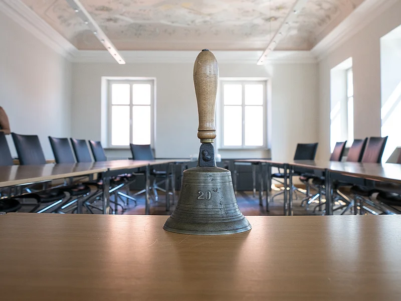 Im Vordergrund eine Handglocke mit Holzgriff auf einem Tisch in einem Sitzungssaal mit bemalter historischer Stuckdecke, die biblische Szenen zeigt und schwarze Stühle entlang des U-förmigen Tisches. Einige Fenster entlang der zwei Wände.