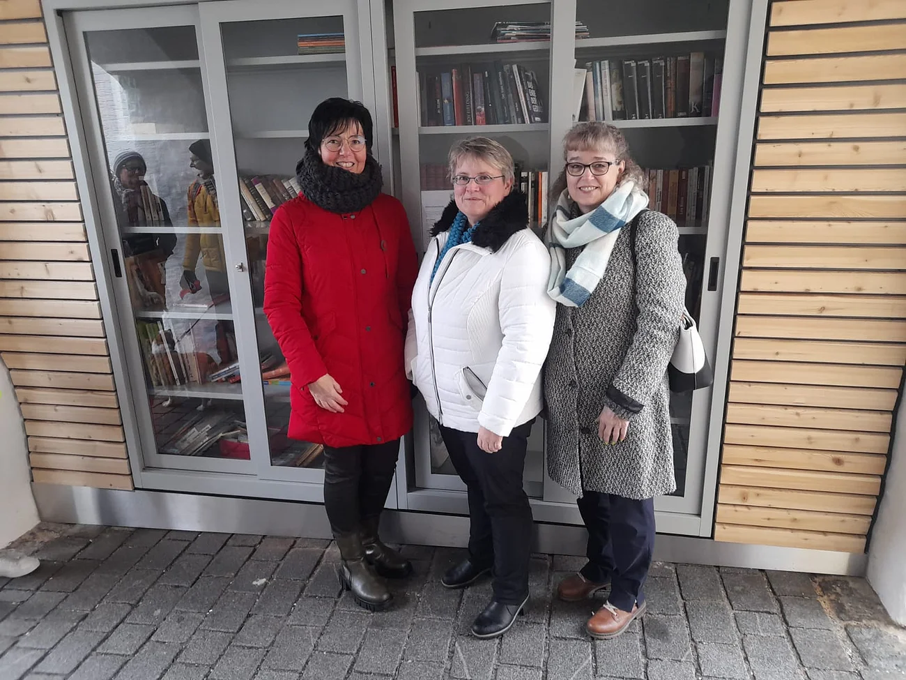 Offizielle Eröffnung des offenen Bücherschrankes Drei Frauen in Winterkleidung stehen vor einem Bücherschrank mit Glastüren links und rechts am Schrank eine Holzfassade.