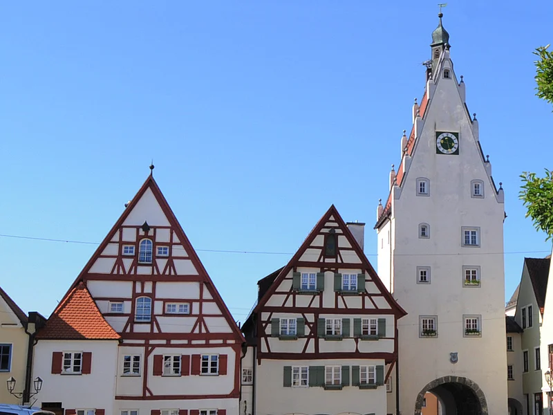 Historische Fachwerkhäuser, ein Torturm mit Uhr in einer Altstadt bei klarem Himmel. Links im Bild parkende Autos auf einem gepflasterten Platz. Rechts im Vordergrund ein Baum mit einer Info-Tafel und einem großen Buchstaben "O" aus Stein. Dahinter eine Informationsvetrine.