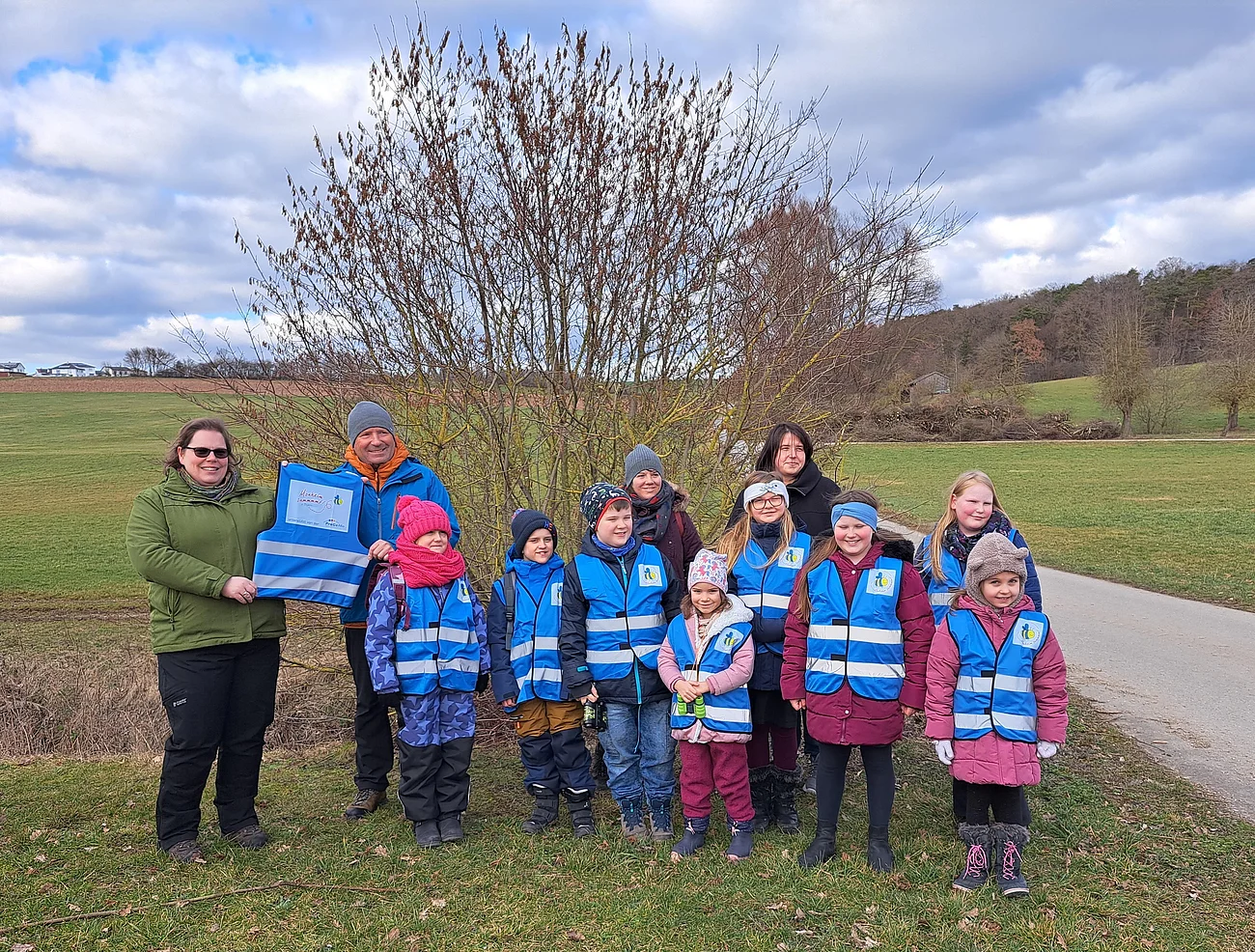 Eine Gruppe von Erwachsenen und Kindern in blauen Westen steht im Freien auf einem Feld vor einem Baum. Zwei erwachsen Personen halten zusammen eine blaue Weste in den Händen. Rechts von ihnen verläuft ein Schotterweg. Im Hintergrund blauer Himmel.