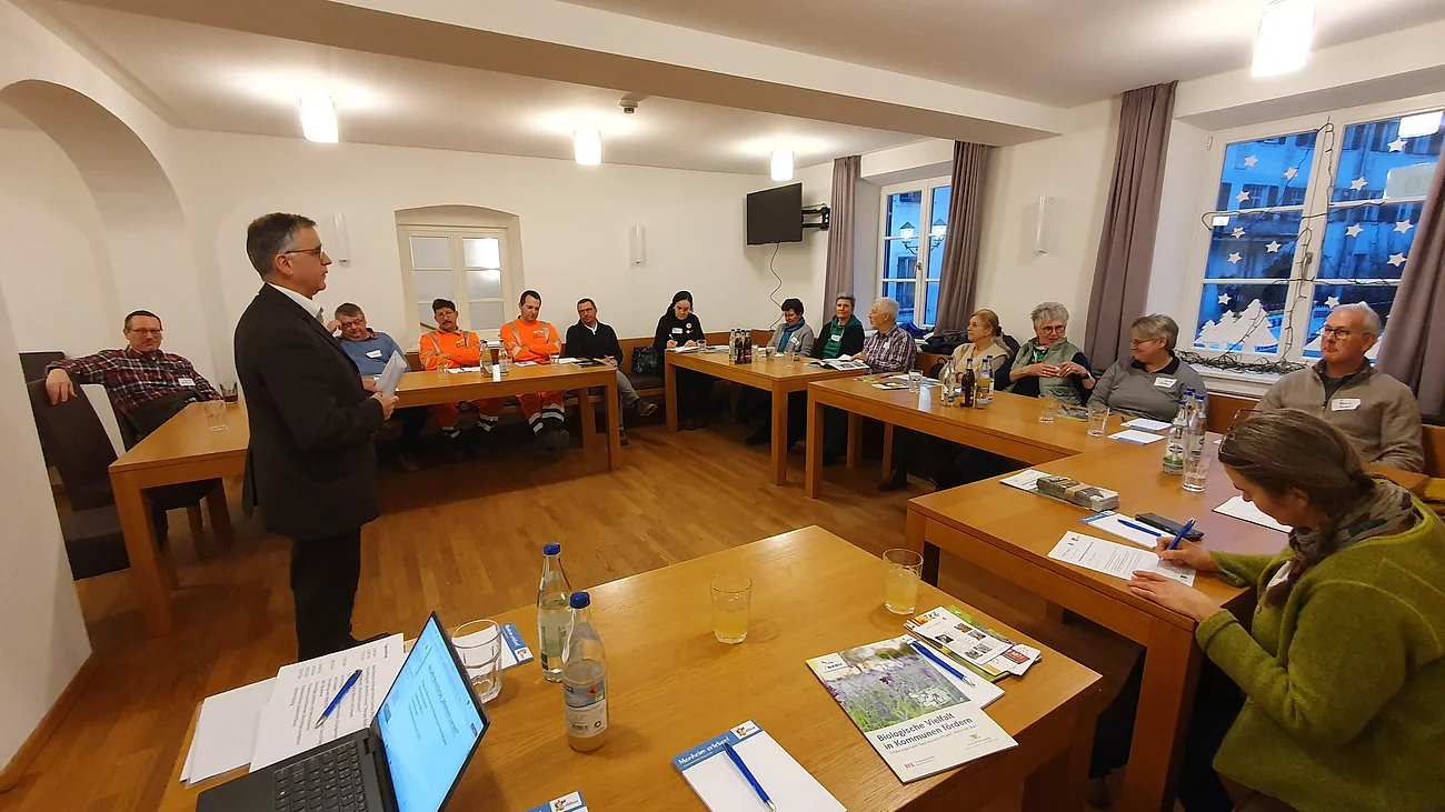 Eine Gruppe von Menschen sitzt in einem Besprechungsraum an Tischen, während eine vorne Person steht und spricht. Tische sind mit Getränken und Unterlagen bedeckt.