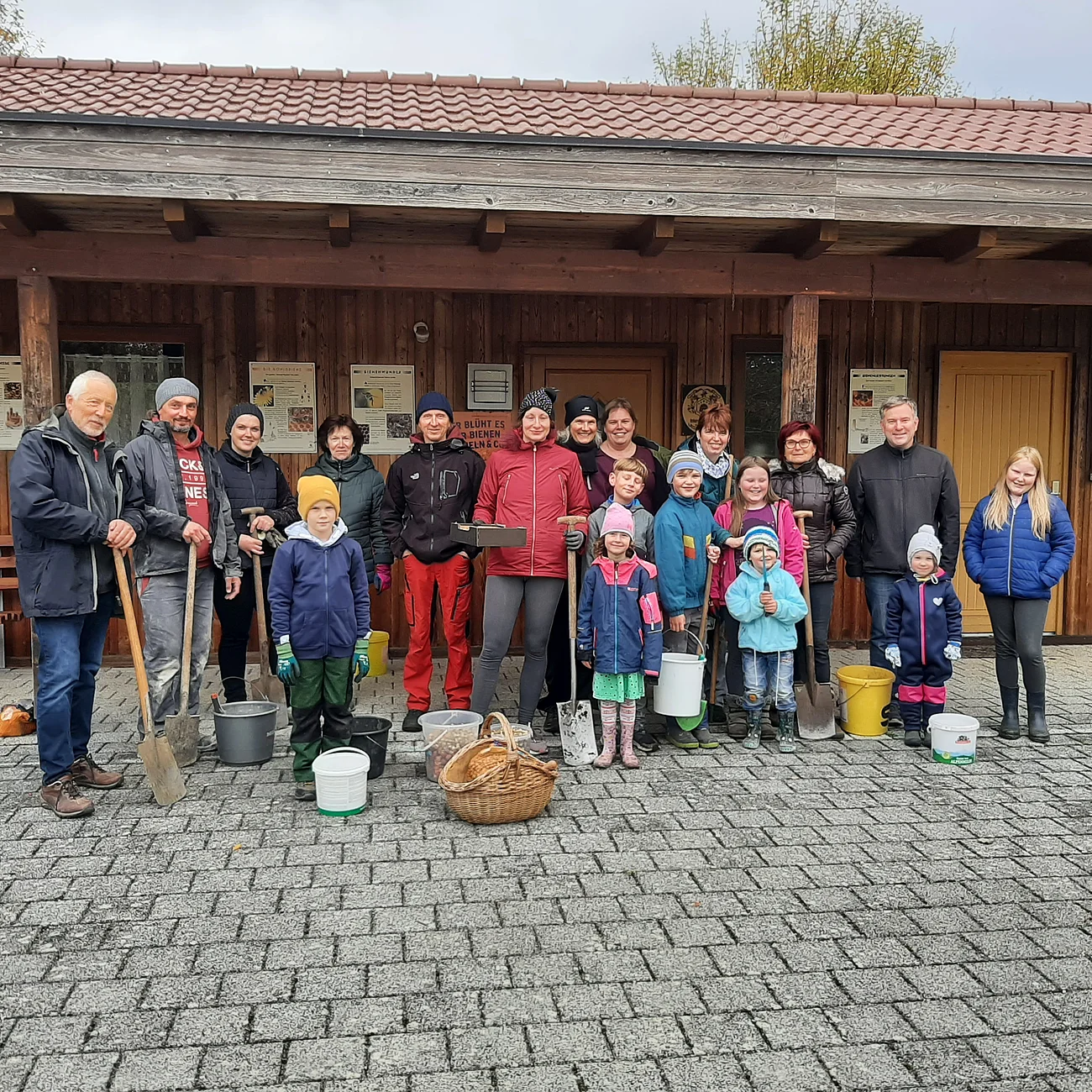Gruppe von Menschen mit Gartenwerkzeugen und Eimern und einem geflochtenen Korb, steht vor einem Holzgebäude auf gepflastertem Boden.