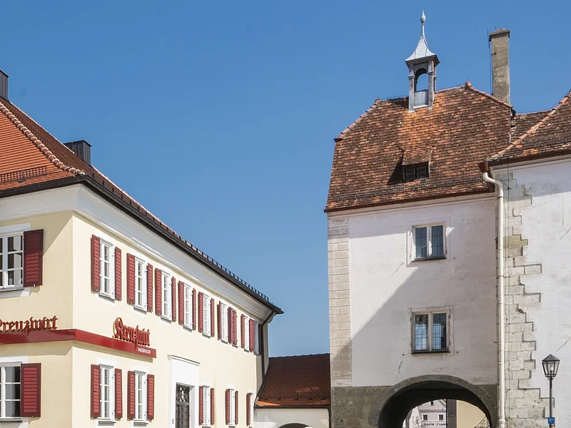 Tourist-Information der Stadt Monheim/Monheimer Alb: ein Fachwerkhaus mit drei Stockwerken, weißen Wänden und braunen Balken. Davor steht ein Schaukasten und zwei Radfahrer fahren auf gepflasterter Straße vorbei. Rechts davon ein gelbes zweistöckiges Gebäude mit roten Fensterläden.
