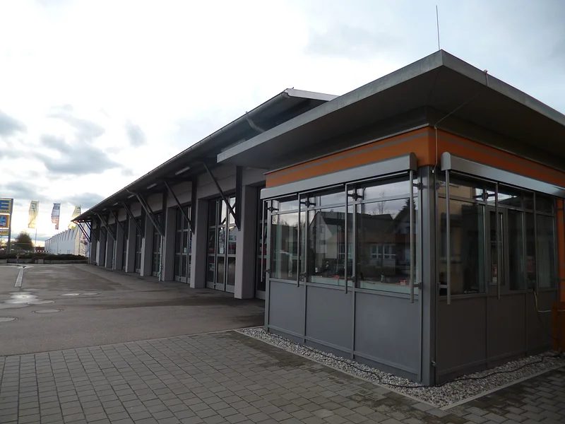 Feuerwehrhaus Monheim Außenansicht eines modernen grauen Feuerwehrhauses mit Glasfront, flachem Dach, vielen großen Toren und gepflastertem Vorplatz bei bewölktem Himmel.
