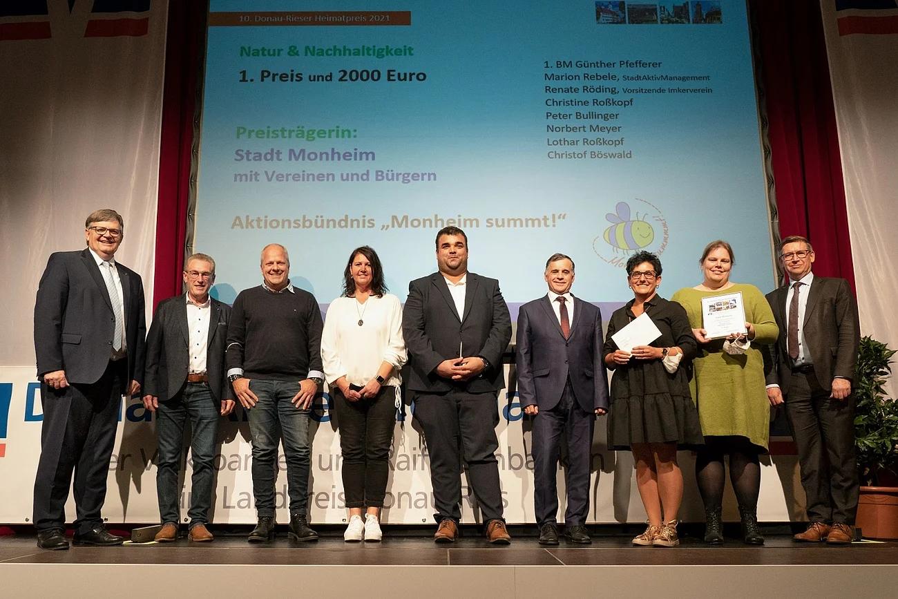 Eine Gruppe von neun Personen steht auf einer Bühne. Im Hintergrund ist eine Leinwand mit Text über einen Preis für Natur und Nachhaltigkeit. Links und recht von der Leinwand ein weißer Vorhang. Zwei Personen halten ein Dokument in den Händen.