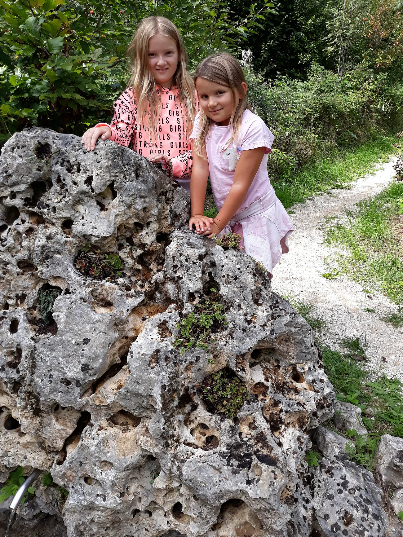 Zwei Mädchen lehnen an einem großen, porösen Felsen neben einem schmalen Weg im Grünen.