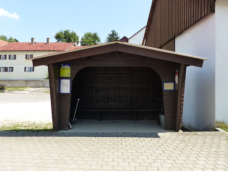 Holzunterstand einer Bushaltestelle auf einem gepflastertem Platz mit Bank, Tretroller und Eimer. An dem Bushaltestellen-Haus zwei Aushänge mit den Fahrplänen und ein grün-blaues Schild mit der Aufschrift "SoMit Bürgerbus" angebracht. Dahinter Häuser, grüne Bäume und blauer Himmel bei Tag.