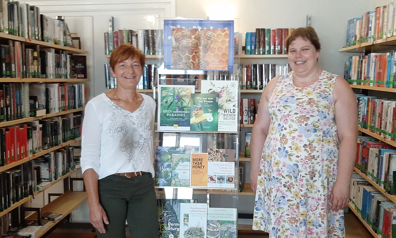 Zwei lächelnde Frauen stehen in einer Bibliothek neben einem Bücherständer mit Fachbüchern über Bienen und Natur.