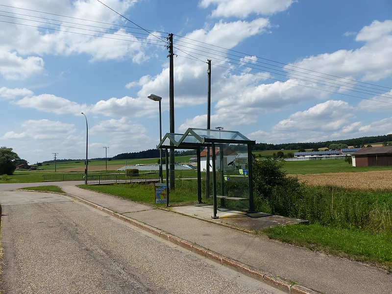 Haltestelle mit Glasunterstand an einer Landstraße, umgeben von Feldern und blauem Himmel mit Wolken. Auf dem Bushäuschen ein Aushang mit dem Fahrplan und ein grün-blauer Sticker mit der Aufschrift "SoMit Bürgerbus" angebracht.