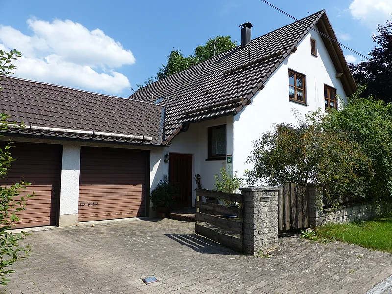Weißes Einfamilienhaus mit braunem Ziegeldach, zwei braunen Garagentoren, gepflastertem Hof und Holzzaun vor grünem Buschwerk. Im Hintergrund blauer Himmel.
