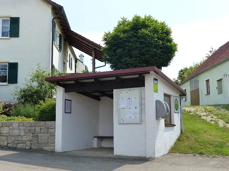 Weißes Buswartehäuschen aus Stein mit einer Sitzbank darin, einem Informationskasten außen am Häuschen, einem Haltestellenschild, einem Fenster, und einem Defibrillator-Kasten. Außerdem ist ein Aushang mit dem Fahrplan und ein grün-blaues Schild mit der Aufschrift "SoMit Bürgerbus" angebracht. Wohnhäuser mit Garten, Grüner Wiese und Baum im Hintergrund.