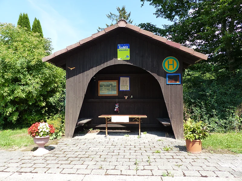 Braunes Holzwartehäuschen darin: zwei Bänke links und rechts, ein Tisch, ein eingerahmtes Bild, der Aushang mit Fahrplan und Dekorationsartikel. Auf dem Bushäuschen ein Bus-Haltestellenschild und ein grün-blaues Schild mit der Aufschrift "SoMit Bürgerbus" außen angebracht. Das Häuschen umgeben von grünen Bäumen und zwei bepflanzten Blumentöpfen.
