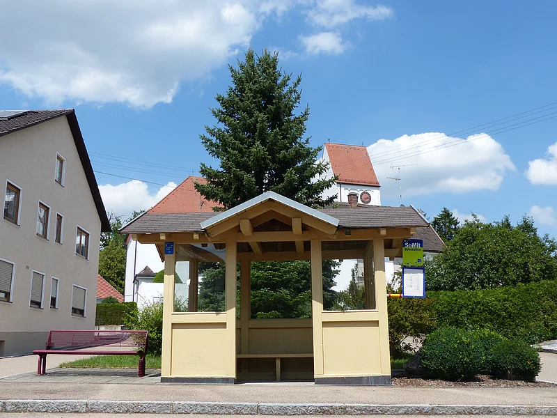 Bushaltestelle mit gelbem Unterstand und braunem Dach, einer roter Bank links daneben, Wohnhaus links und Kirche mit Turmuhr im Hintergrund. Bushaltestelle umgeben von grünen Hecken und einem großen Baum bei klarem Himmel. Auf dem Bushäuschen ein Aushang mit dem Fahrplan und ein grün-blaues Schild mit der Aufschrift "SoMit Bürgerbus" angebracht.