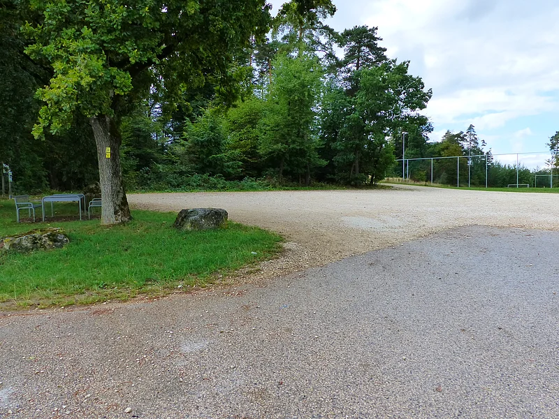 Parkplatz mit Kiesfläche, umgeben von Bäumen, links im Bild unter dem Baum eine Sitzgruppe aus Metall. Im Hintergrund eine Absperrung zum Fußballfeld unter bewölktem Himmel.