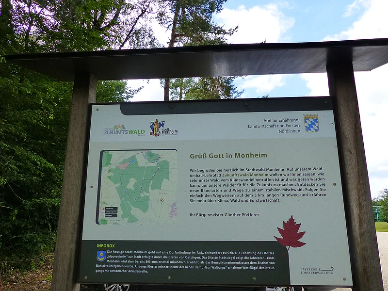 Ein überdachtes Informationsschild aus Holz im Wald mit Text über den Stadtwald Monheim und einer Karte des Waldgebiets.