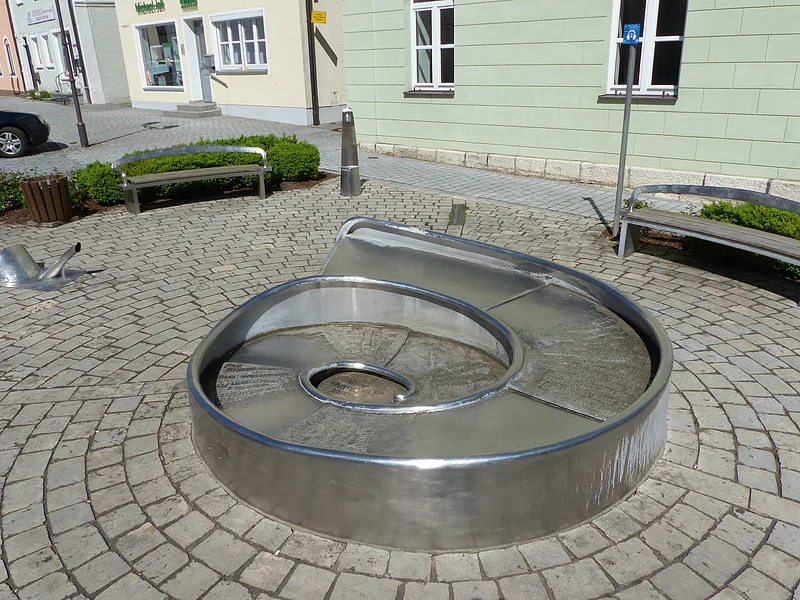 Juraschneck-Brunnen: ein Brunnen aus Metall in Form eines Fossils einer Schnecke, mit eingravierten Texten auf der Oberfläche. Außenrum ein gepflastertem Platz mit zwei Sitzbänken. Im Hintergrund farbige Häuser und eine kleine grüne Hecke an den Bänken entlang.