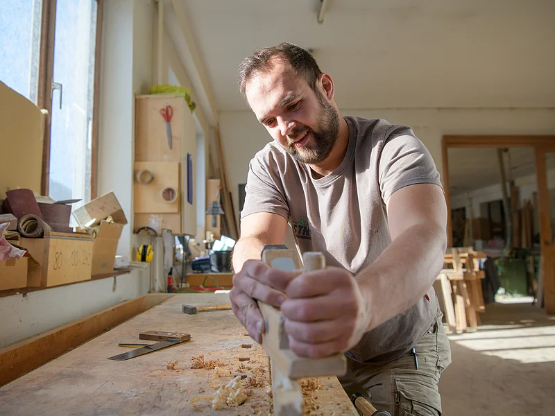 Schreinerei & Küchenstudio Gerstner - Werkstatt Mann hobelt Holz auf Werkbank in heller Werkstatt mit Werkzeug vor einem Fenster.