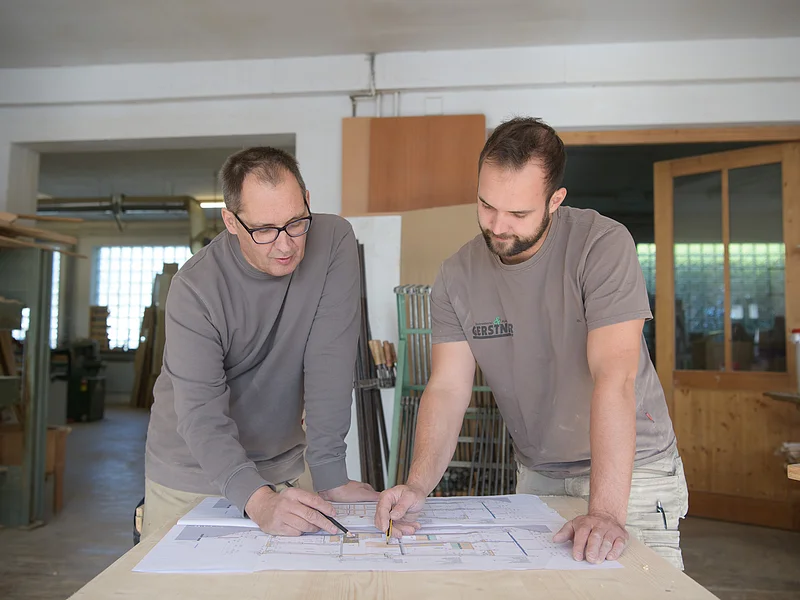 Schreinerei & Küchenstudio Gerstner - Planung Zwei Männer in Werkstatt besprechen Baupläne auf einem Tisch, einer zeigt mit Stift darauf.