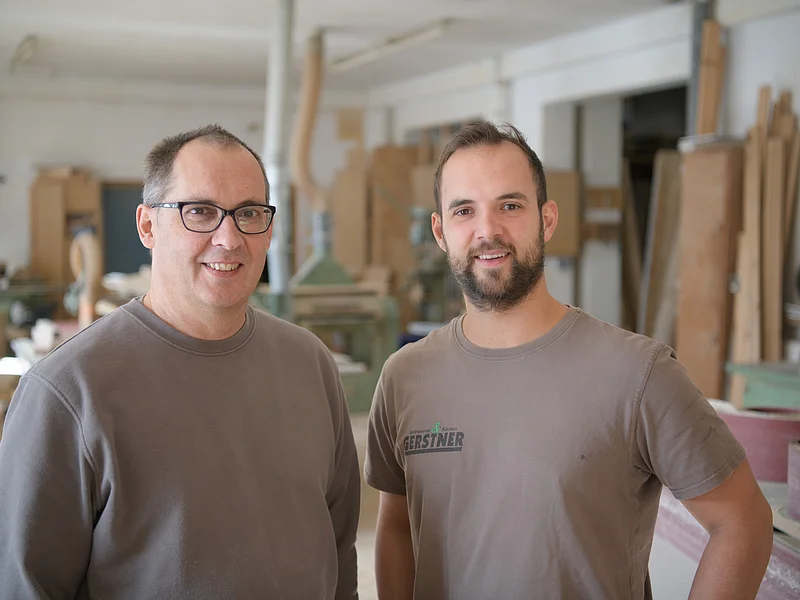 Schreinerei & Küchenstudio Gerstner - Team Zwei Männer in grauen Oberteilen stehen in einer Werkstatt mit Holzbearbeitungsmaschinen.