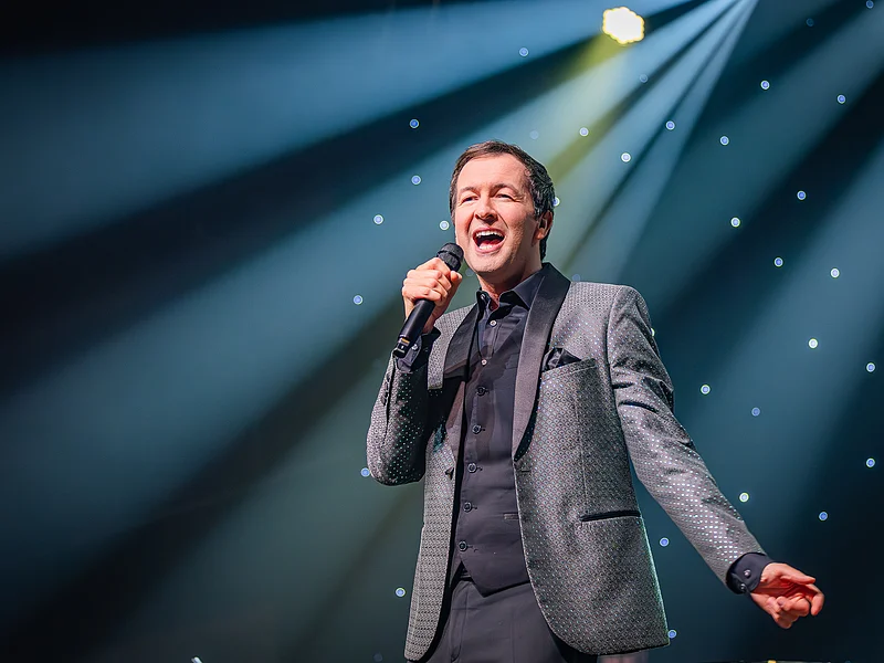 Udo Jürgens - Das Konzert mit Alex Parker & Special Guest Mann im Anzug singt mit Mikrofon auf Bühne vor beleuchtetem Hintergrund mit Sternenlicht.
Udo Jürgens - Das Konzert mit Alex Parker & Special Guest am 20.02.2027 in der Monheimer Stadthalle.