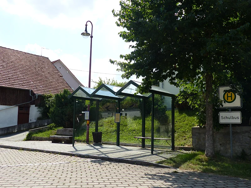 Haltestelle mit Glasunterstand an einem Gehweg vor einer gepflasterten Straße, umgeben von Bäumen, grüner Wiese und einer Straßenlaterne dahinter. Links davon ein weißes Gebäude und eine Holzsitzbank bei blauem Himmel. Rechts im Bild ein grün-gelbes Bushaltestellenschild mit einem zweiten Schild darunter mit der Aufschrift "Schulbus".