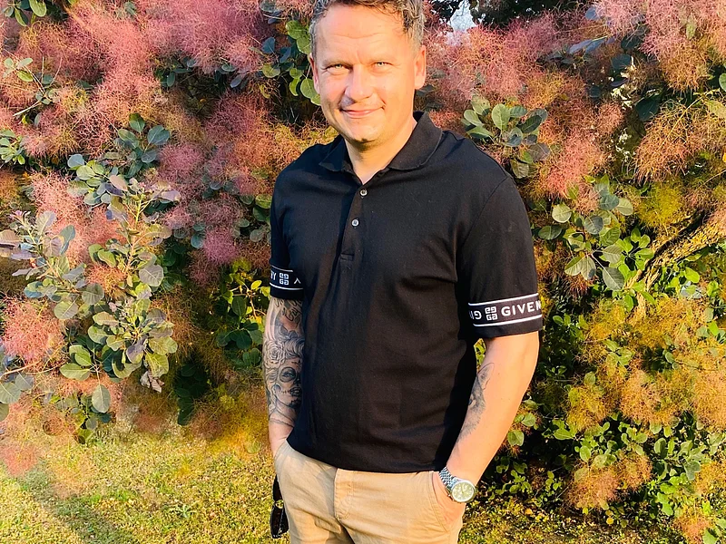 Mann mit schwarzem Poloshirt und beigen Shorts steht auf Rasen vor buschigem Baum mit rosa Blüten.