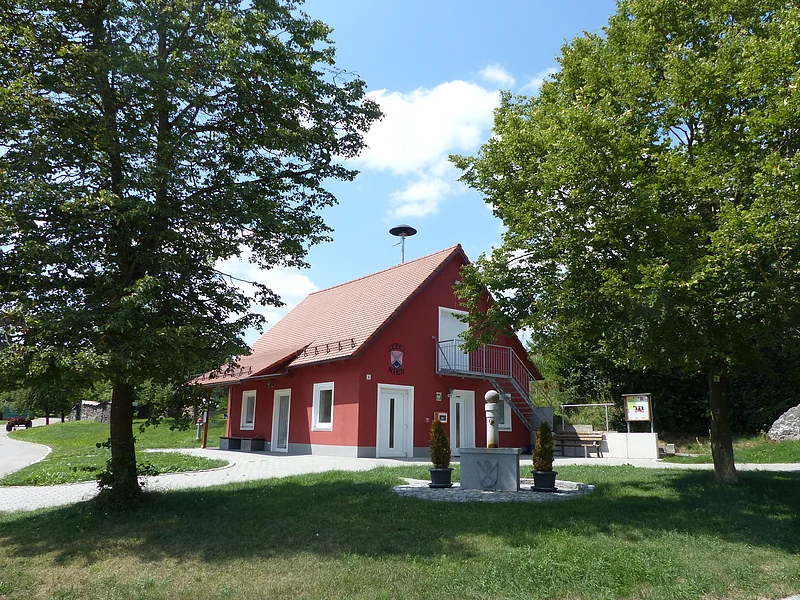 Rotes Gebäude mit rotem Dach, weißen Tür- und Fensterrahmen, einem kleinen Vordach und einer Metalltreppe in den ersten Stock, umgeben von Bäumen und Rasen unter blauem Himmel mit Wolken. Vor dem Gebäude ein Brunnen mit zwei Pflanztöpfen und an der Eingangstür hängt ein Defibrillator in einem orangen Kasten an der Wand.
