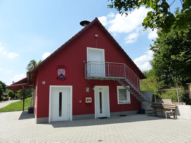 Rotes Gebäude mit rotem Dach, weißen Tür- und Fensterrahmen, einem kleinen Vordach und einer Metalltreppe in den ersten Stock, umgeben von Bäumen und Rasen unter blauem Himmel mit Wolken. Vor dem Gebäude ein gepflasterter Vorhof mit einer Holz-Sitzbank und an der Eingangstür hängt ein Defibrillator in einem orangen Kasten an der Wand.