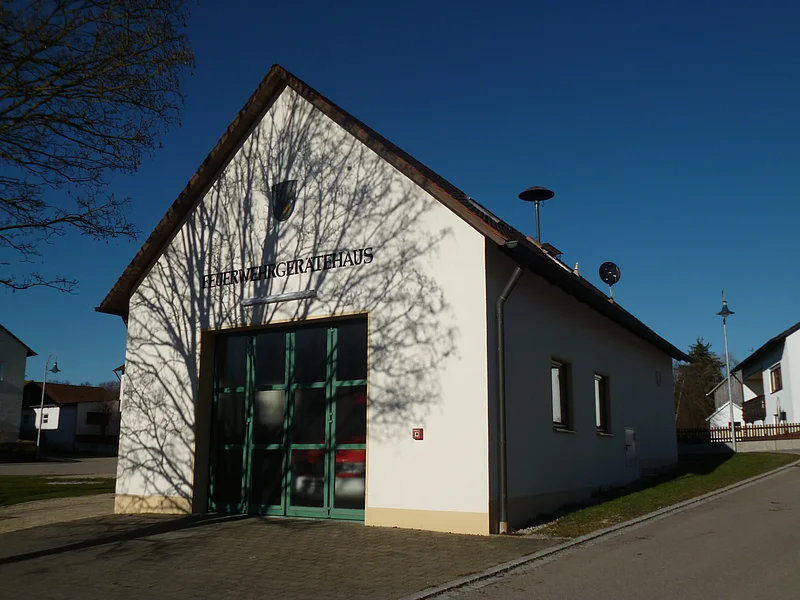 Feuerwehrhaus mit einem großen grünen Tor, Fenstern und braunem Dach an einer Straße. Auf dem weißen Gebäude die Aufschrift "Feuerwehrgerätehaus" und Schatten eines Baumes auf der Fassade, bei klarem Himmel. Links im Eck des Bildes ein Baum zu sehen.