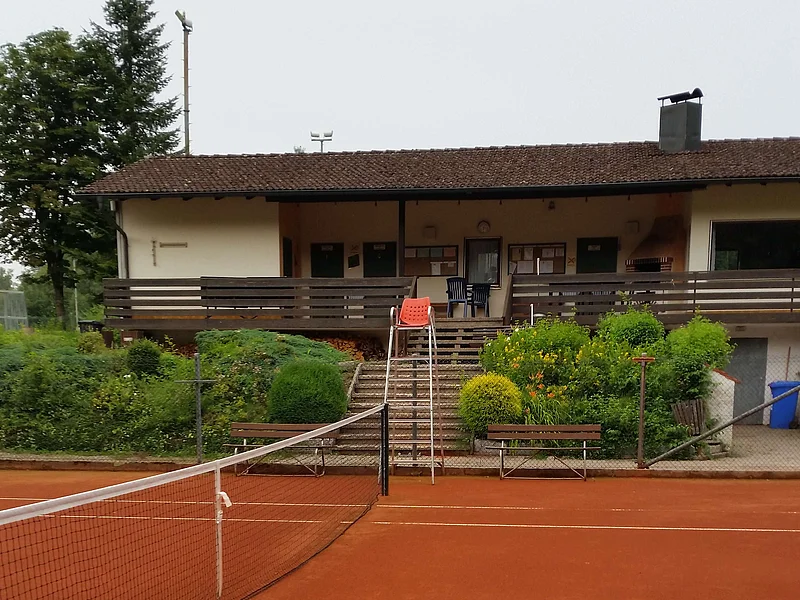 Tennisanlage Monheim Tennisplatz mit rotem Sandbelag, einem Netz in der Mitte, dahinter ein Gebäude mit braunem Dach, einer Terrasse und Treppe, umgeben von Büschen und Bäumen.