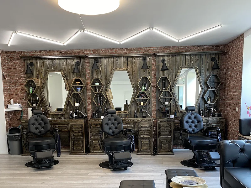 Friseursalon mit drei schwarzen Friseurstühlen vor dunkelbraunen Holzspiegeln und Backsteinwand. Rechts im Bild kleiner Ausschnitt vom Sitzbereich mit schwarzen Möbeln. An der Decke moderne Leuchte.