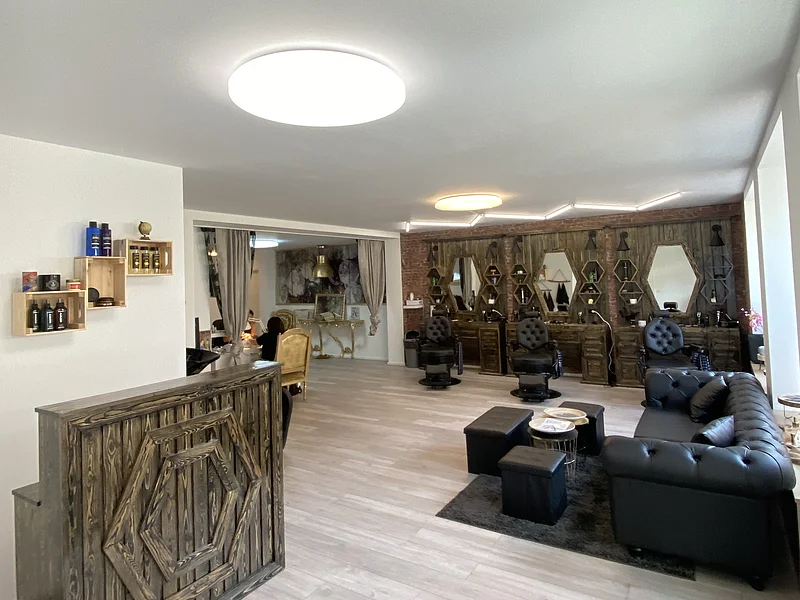 Friseursalon mit dunkelbraunen Holzmöbeln, schwarzen Friseurstühlen, schwarzem Sofa rechts im Bild und großen Spiegeln an der Wand. Links im Bild im Vordergrund eine Empfangstheke in Holzoptik auf hellem Bodenbelag.