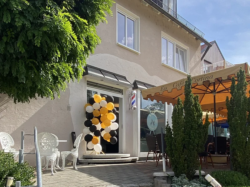 Außentreppe zu einem Eingang, daneben ein Eis-Café. An der Tür hängen gelb-schwarz-weiße Luftballons. Rechts davon der Sitzbereich des Eis-Cafés mit einem orangen Sonnenschirm. Hinter dem Gebäude ein strahlend blauer Himmel.