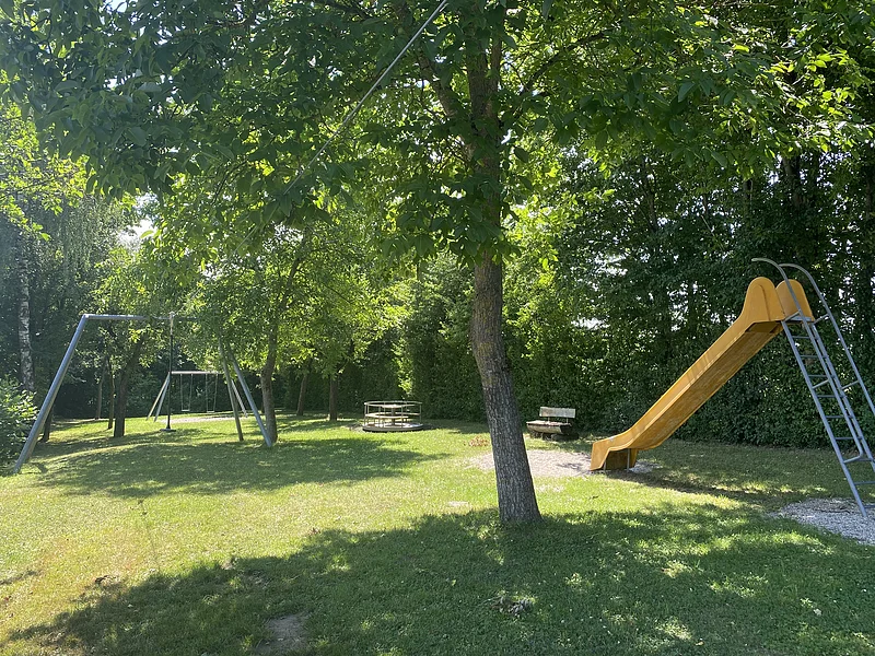 Spielplatz mit Rutsche, Schaukel, Karussell und Bank unter Bäumen auf Grasfläche bei Sonnenschein.