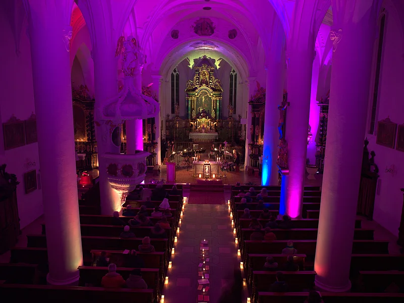 Innenansicht einer Kirche mit violetter Beleuchtung, Kerzen entlang des Gangs und Menschen auf den Bänken. Vor dem Altar spielt eine Musik-Kapelle.