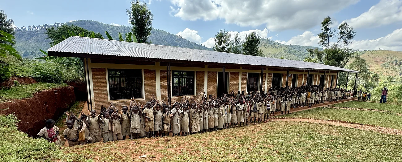Dunkelhäutige Schulkinder in Uniform stehen vor einem langen Backsteingebäude - einer Schule - in hügeliger Landschaft unter bewölktem Himmel. Im Hintergrund sind Bäume zu sehen.