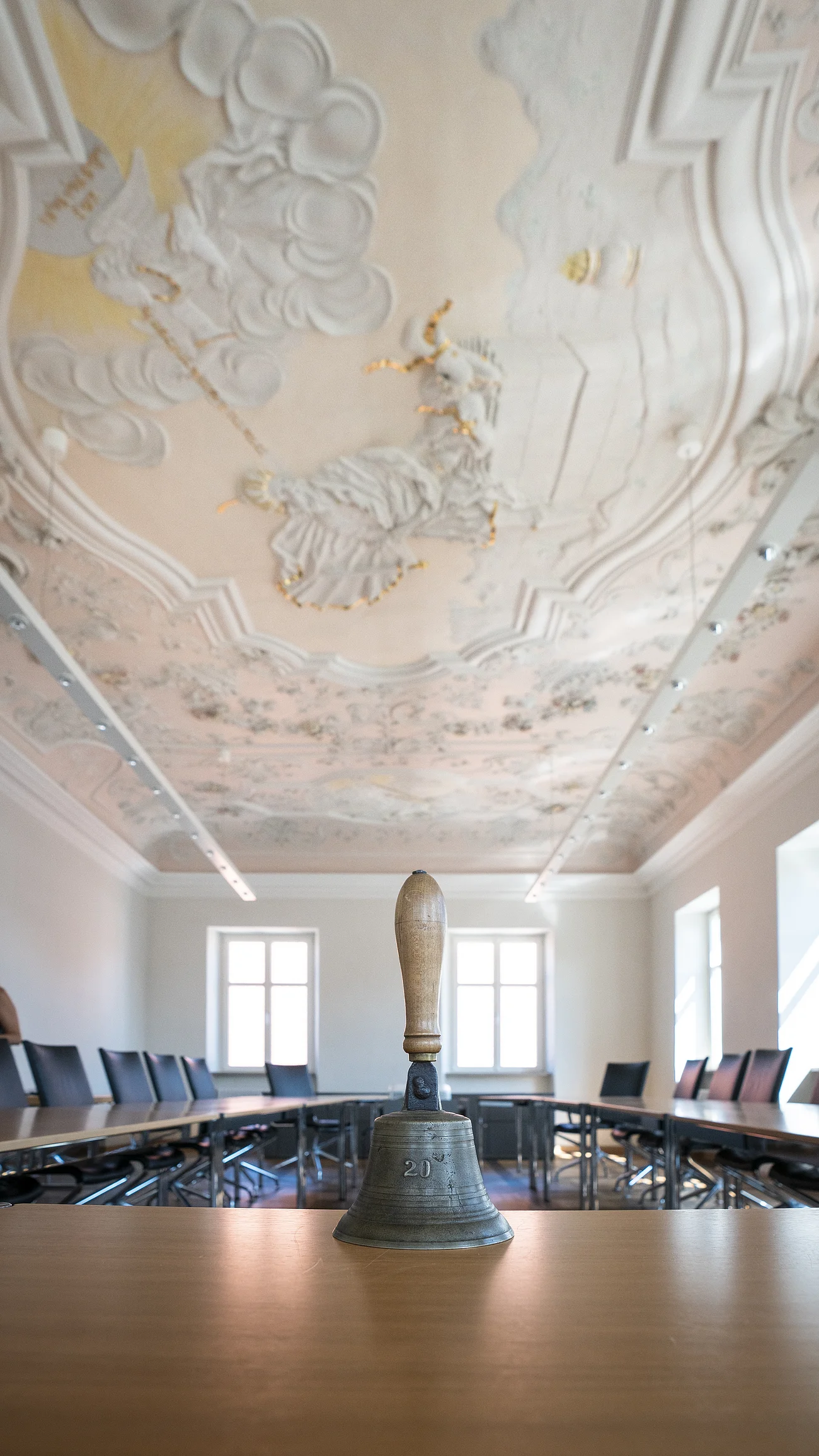 Sitzungssaal Rathaus Im Vordergrund eine Handglocke mit Holzgriff auf einem Tisch in einem Sitzungssaal mit bemalter historischer Stuckdecke, die biblische Szenen zeigt und schwarze Stühle entlang des U-förmigen Tisches. Einige Fenster entlang der zwei Wände.