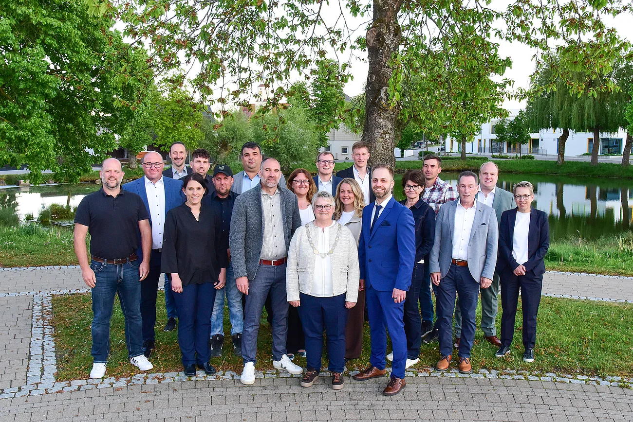 Gruppenfoto von 18 Personen vor einem Teich mit Bäumen und Gebäuden im Hintergrund. Stadtrat Mitglieder der Stadt Monheim - Stand Mai 2026